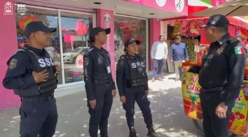 A group of four police officers, three women and one man, engage in conversation outside a vibrant pink shop adorned with colorful decorations and a street vendor cart nearby.$# CAPTION