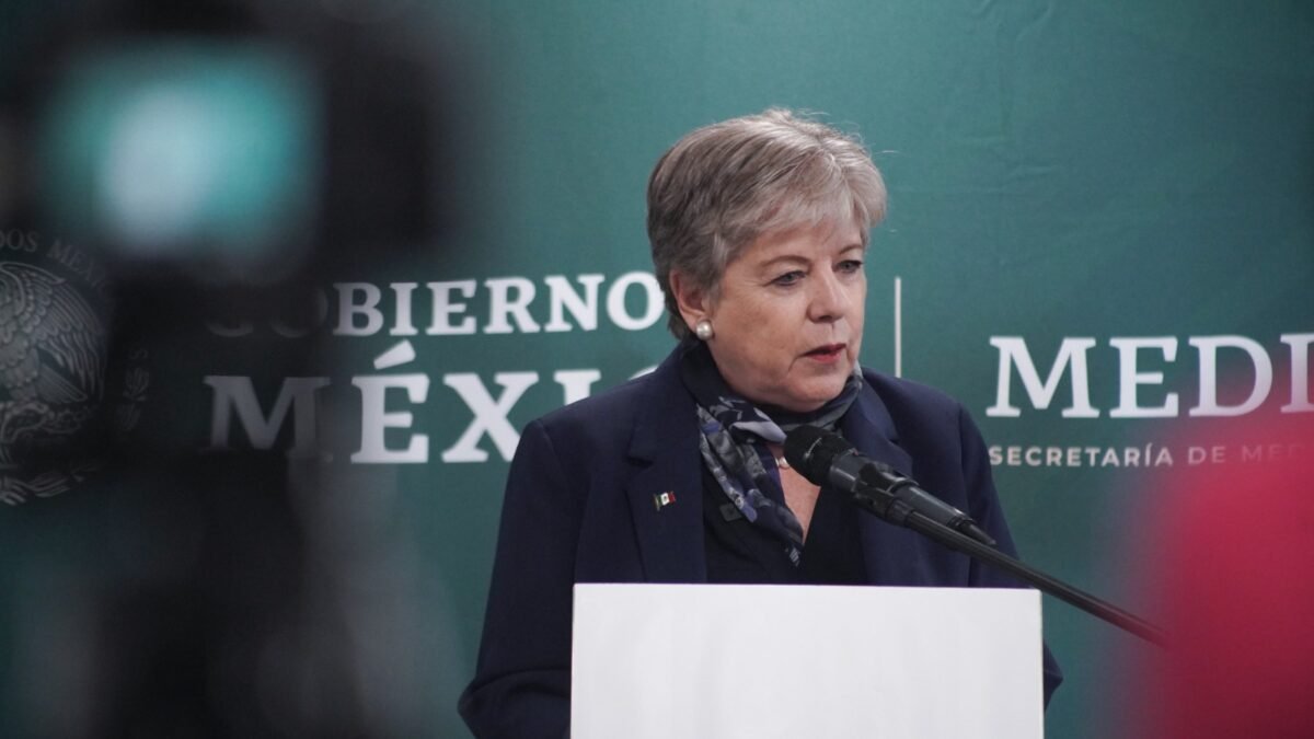 A woman speaking at a podium during a press conference, with a green backdrop featuring the Mexican government insignia and the word "MEDIOS."