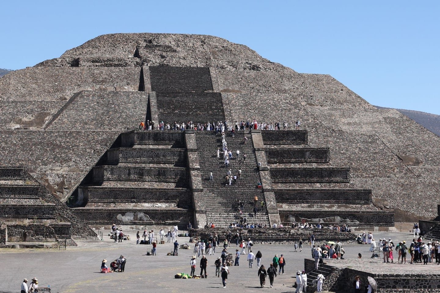 The Pyramid of the Sun, a large Mesoamerican pyramid, with visitors ascending the steps and exploring the base. Clear blue sky above.