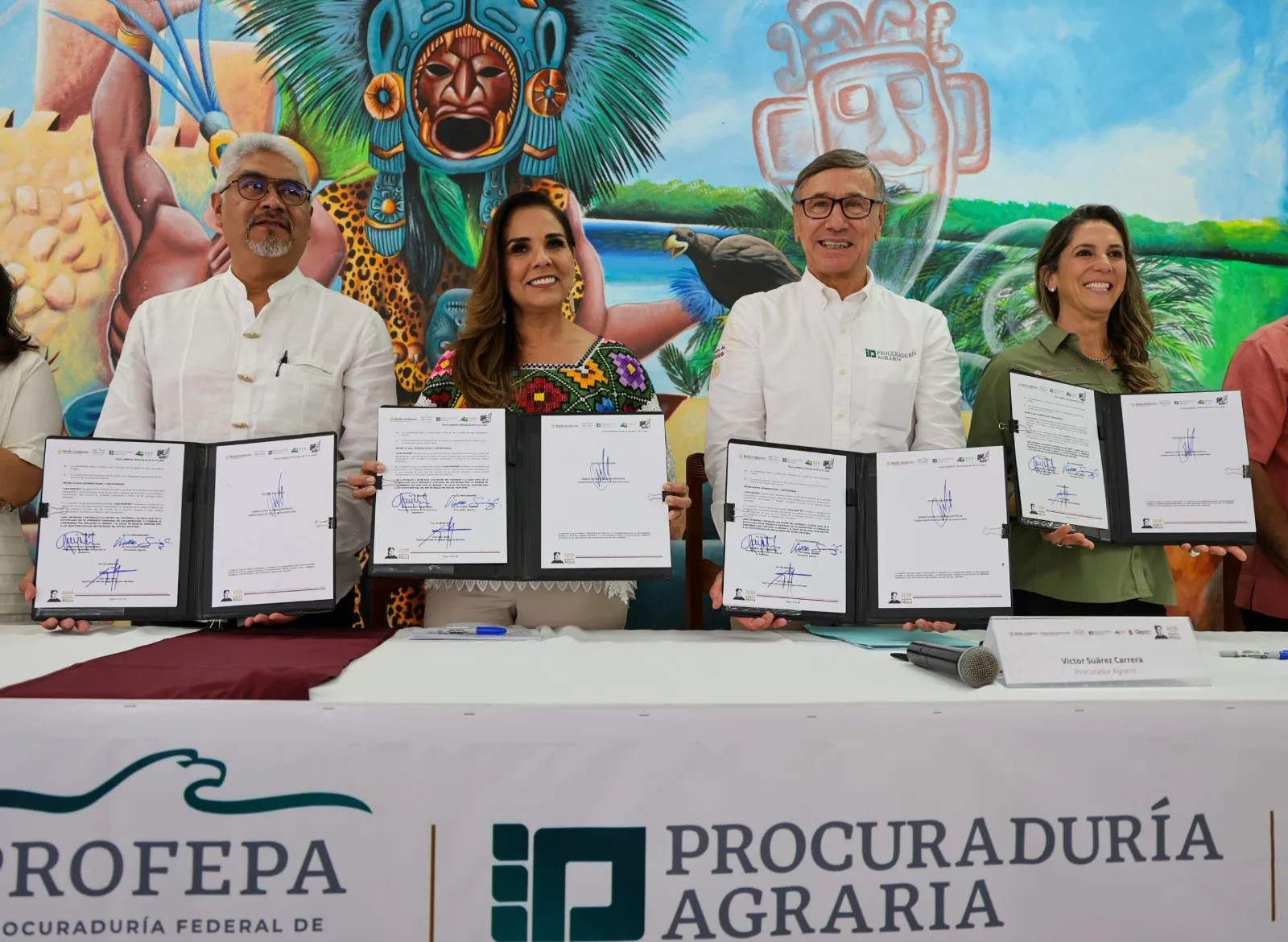Officials standing at a table holding signed documents in front of a colorful mural.$# CAPTION