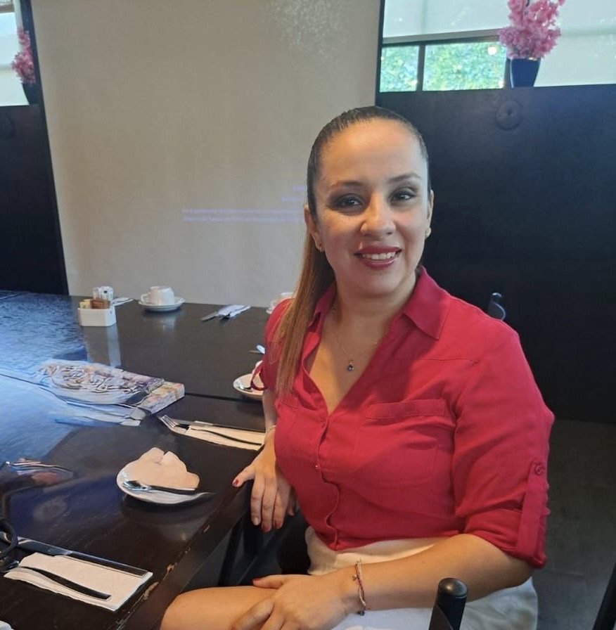 A woman in a red shirt sits at a dining table, smiling. She has long hair and is placed against a backdrop of a restaurant interior with soft lighting and floral decorations.$# CAPTION