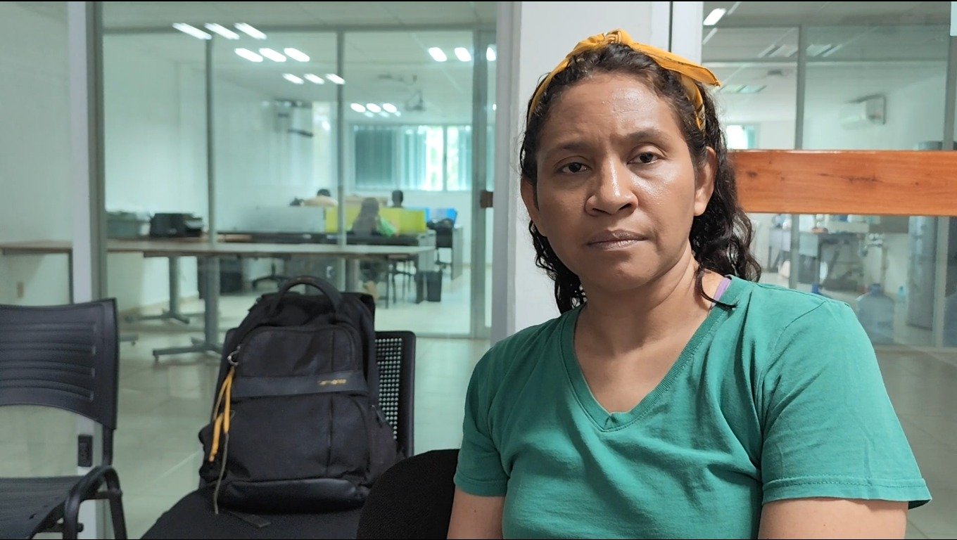 A woman with curly hair wearing a green shirt and a yellow headband, sitting in an office setting with a backpack nearby and people working in the background