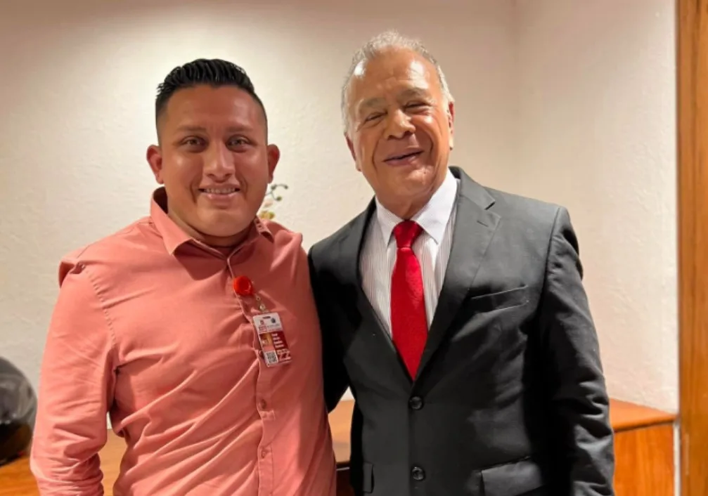Two men smiling and posing for a photo in a professional environment, one in a light pink shirt and the other in a suit with a red tie.$# CAPTION