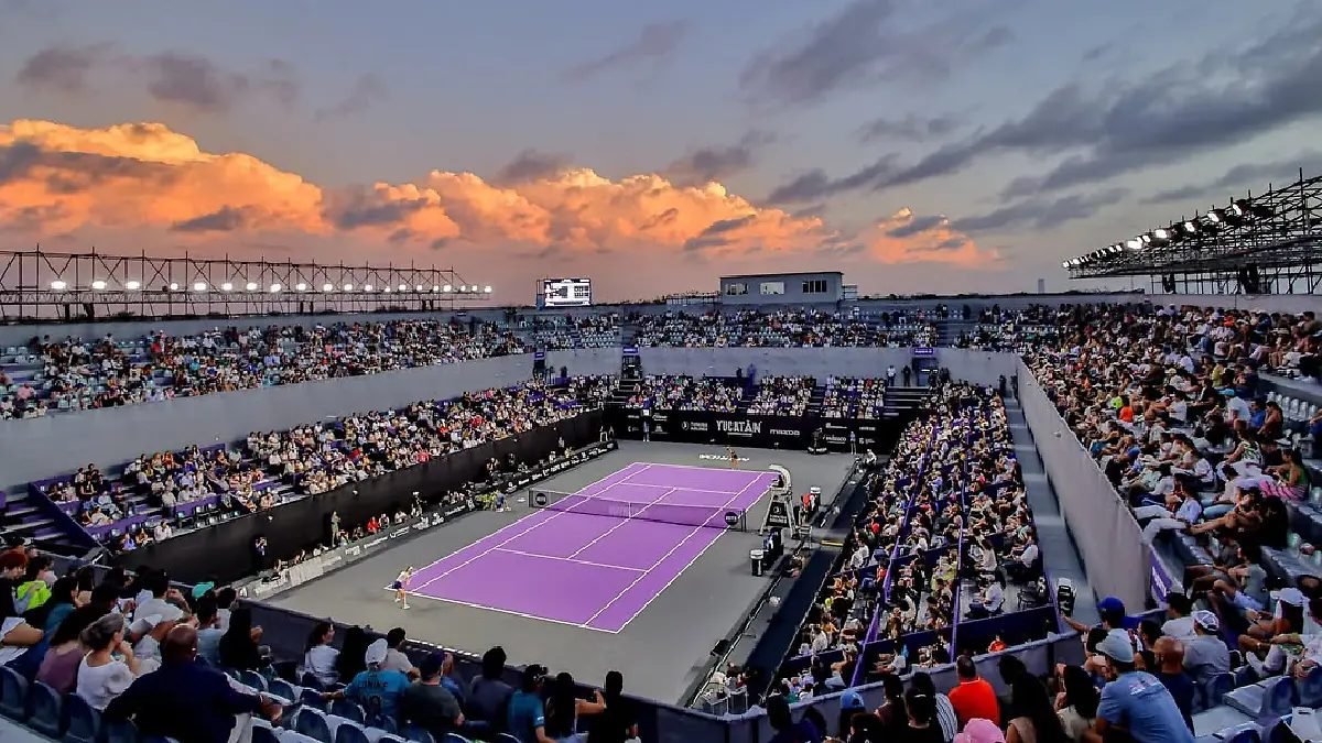 A bustling tennis stadium filled with spectators under a colorful sky at sunset, featuring a purple tennis court.$# CAPTION