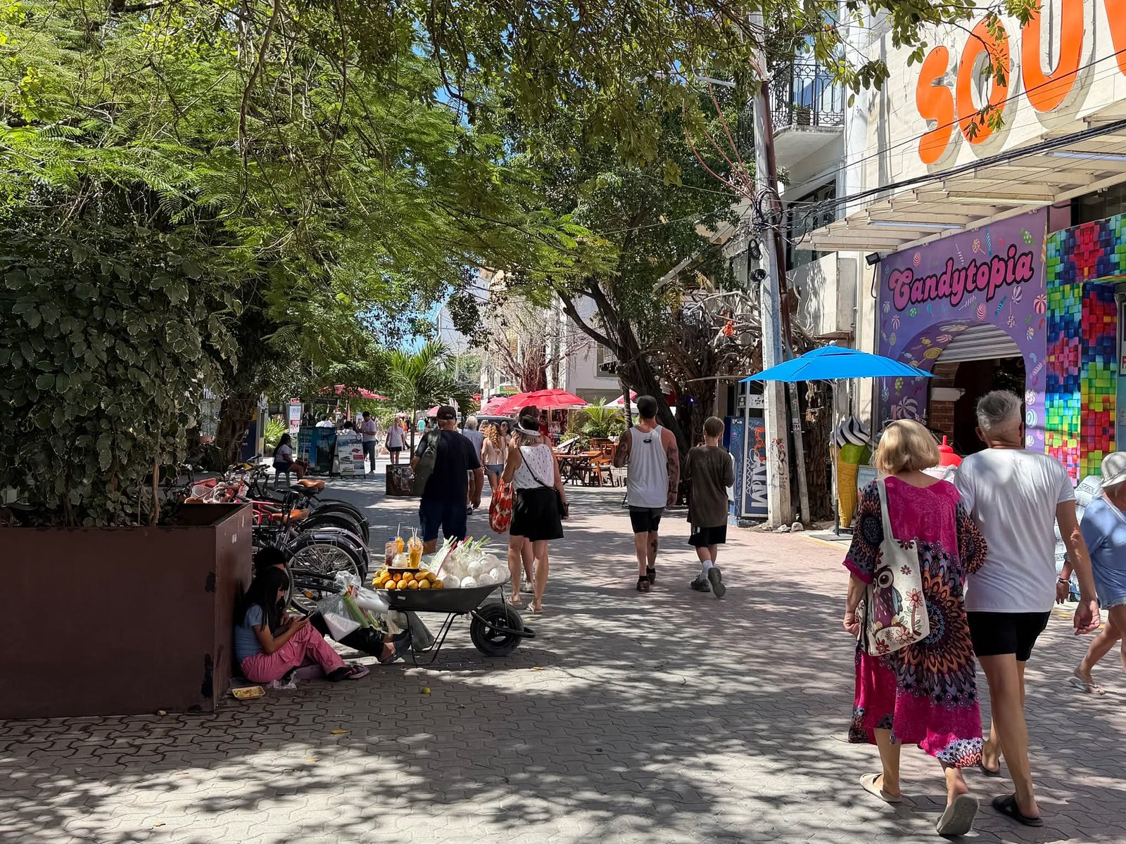 A lively street filled with people walking, shops with colorful umbrellas, and a vendor selling fruits from a wheelbarrow.$# CAPTION