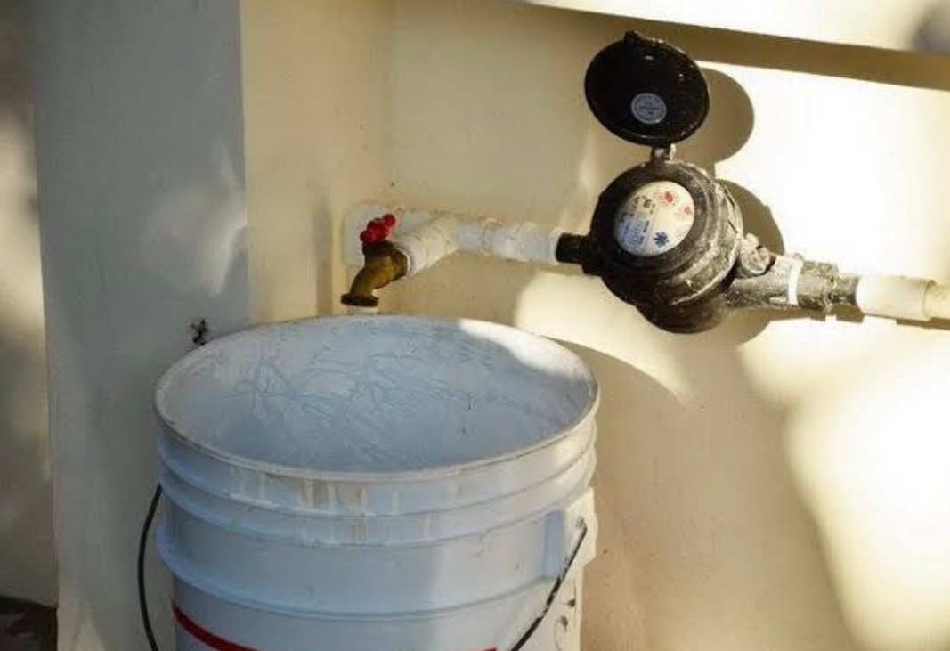 A water meter connected to a pipe leading to a bucket in a residential setting. The bucket is white and has a red stripe, and there is a faucet attached to the pipe.$#$ CAPTION