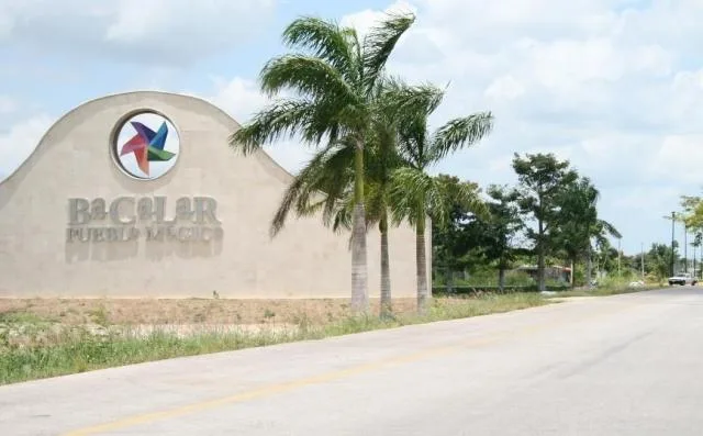 A sign welcoming visitors to Bacalar, Mexico, surrounded by greenery and palm trees, along a road under a blue sky with clouds.$# CAPTION