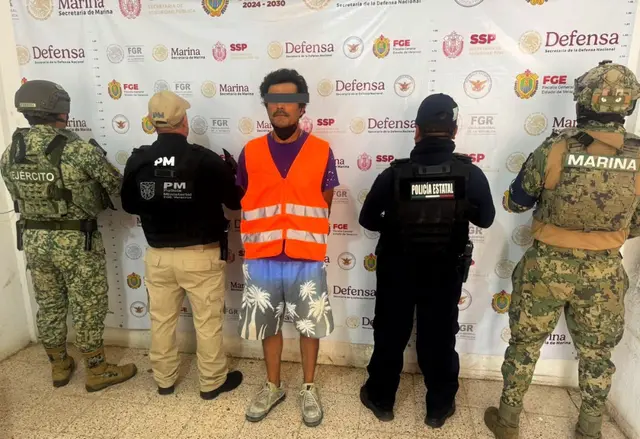 A man in a bright vest stands with his hands behind his back in front of law enforcement officers against a backdrop featuring various governmental logos and insignias.$#$ CAPTION