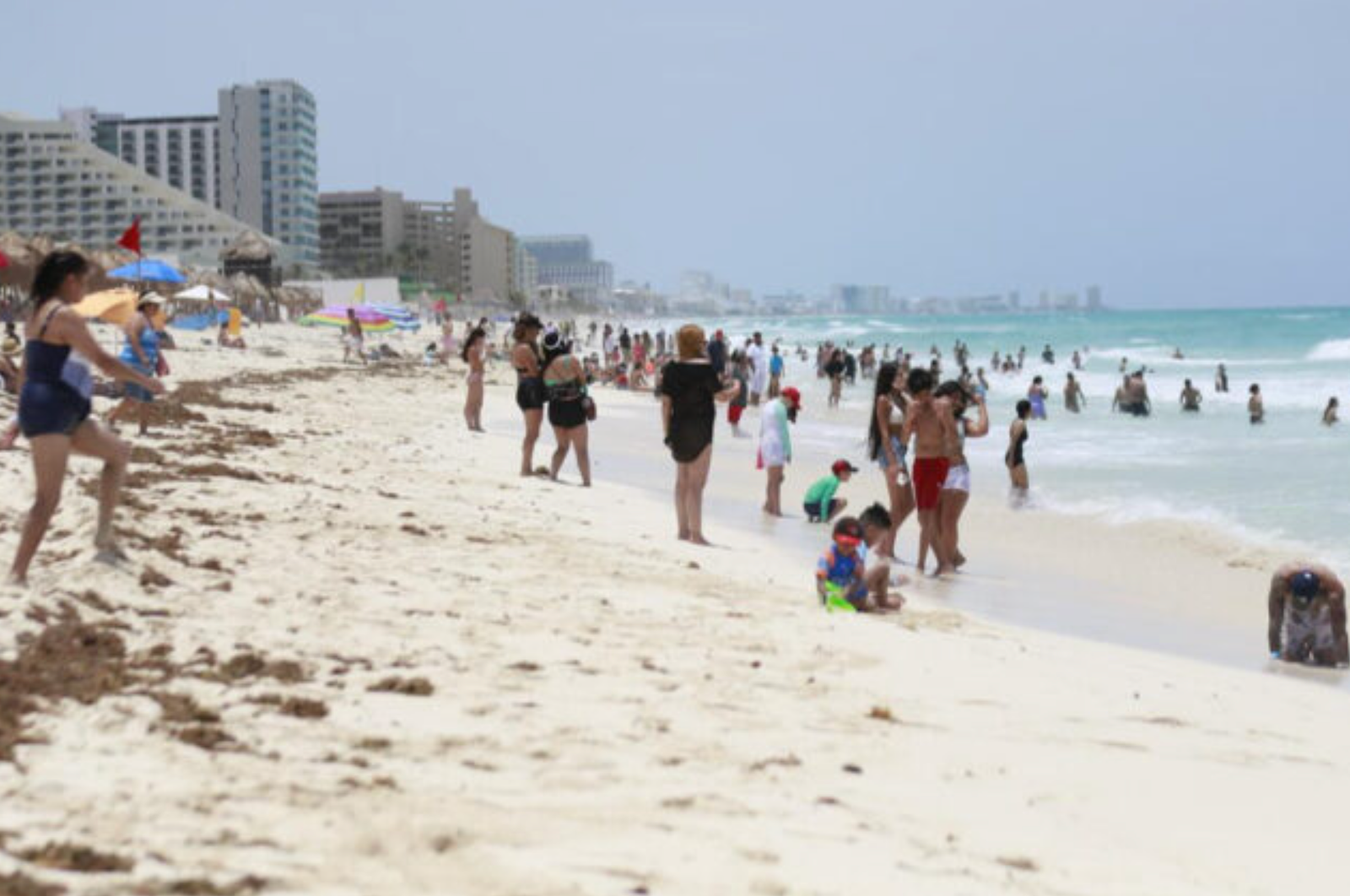 People enjoying a sunny day at the beach in Cancun, with umbrellas and children playing in the sand and water.$#$ CAPTION