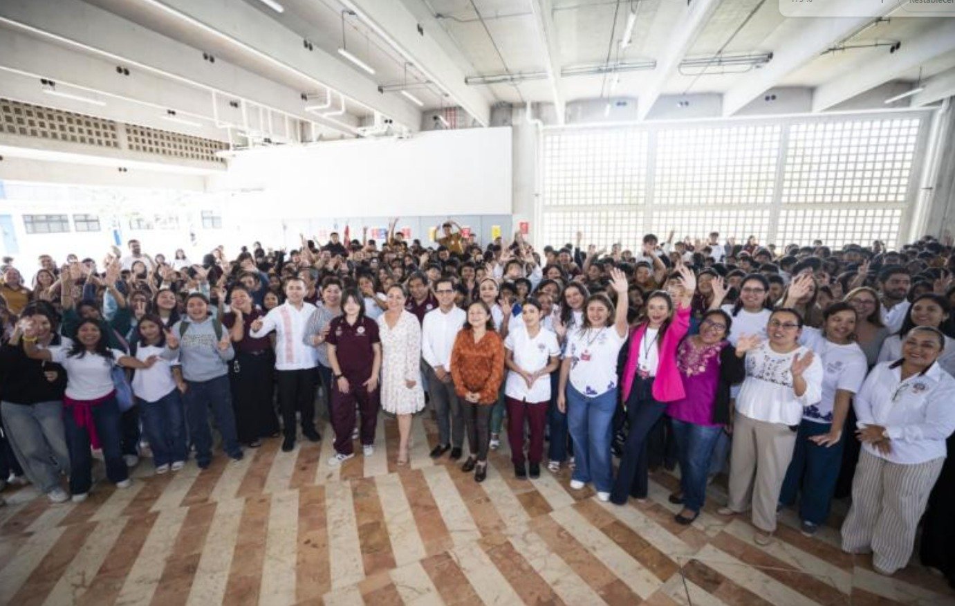A large group of people gathered in a bright indoor space, many smiling and waving at the camera, with some individuals in front dressed in formal attire. A diverse audience is visible in the background, creating a lively atmosphere.