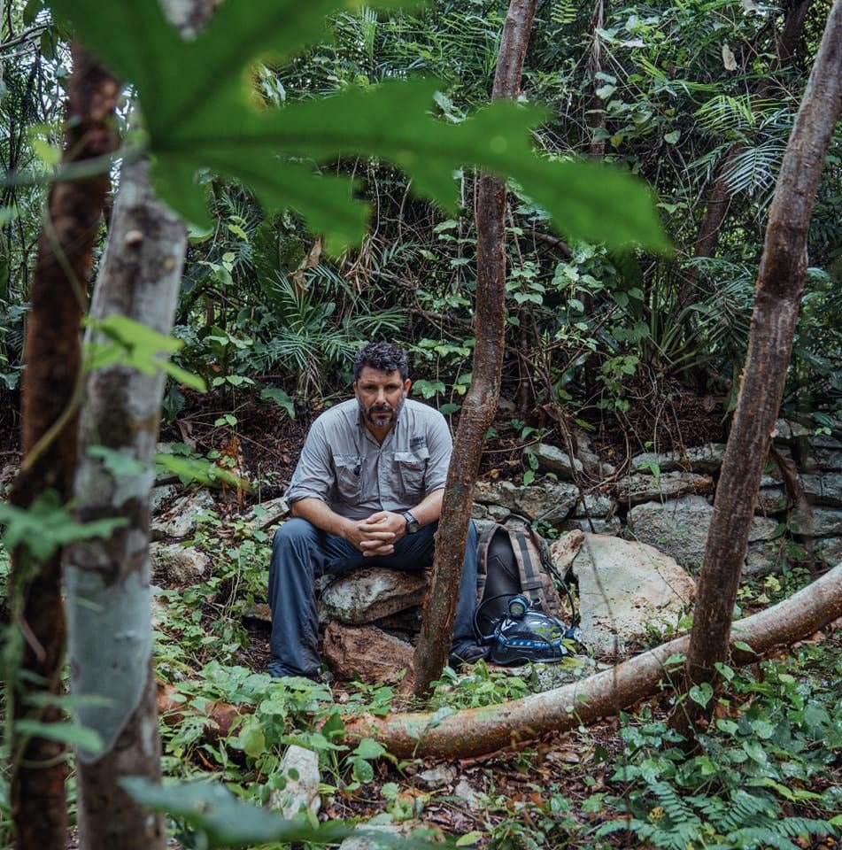A man sits on a rock surrounded by lush greenery in a forest, with a backpack nearby.$# CAPTION