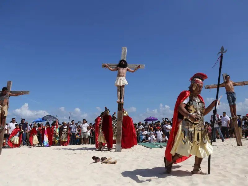 A dramatic reenactment of the crucifixion of Jesus takes place on a beach, with actors on crosses and a crowd watching in the background.$#$ CAPTION