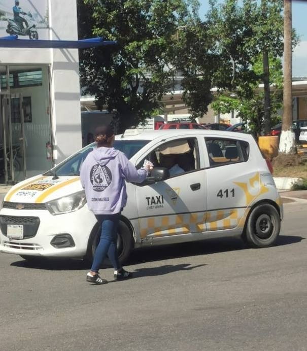 A woman wearing a purple hoodie hands something to the driver of a white taxi in a city street setting. A shopping center can be seen in the background.