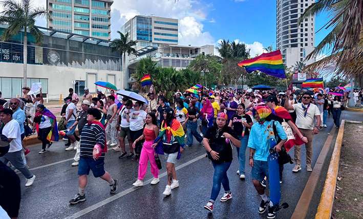 A large crowd of people holding rainbow flags and banners marches along a street in Cancún during the Pride parade.