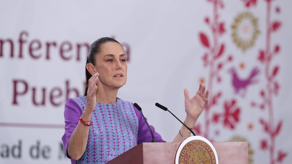 A woman speaking at a podium during a conference, gesturing with her hands, with a decorative background featuring floral designs and text in Spanish.$# CAPTION