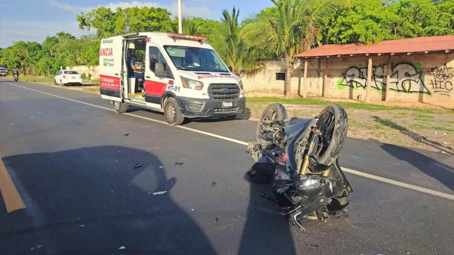A white Chevrolet Aveo with rear-left damage and a motorcycle on the ground on a highway in Tulum
