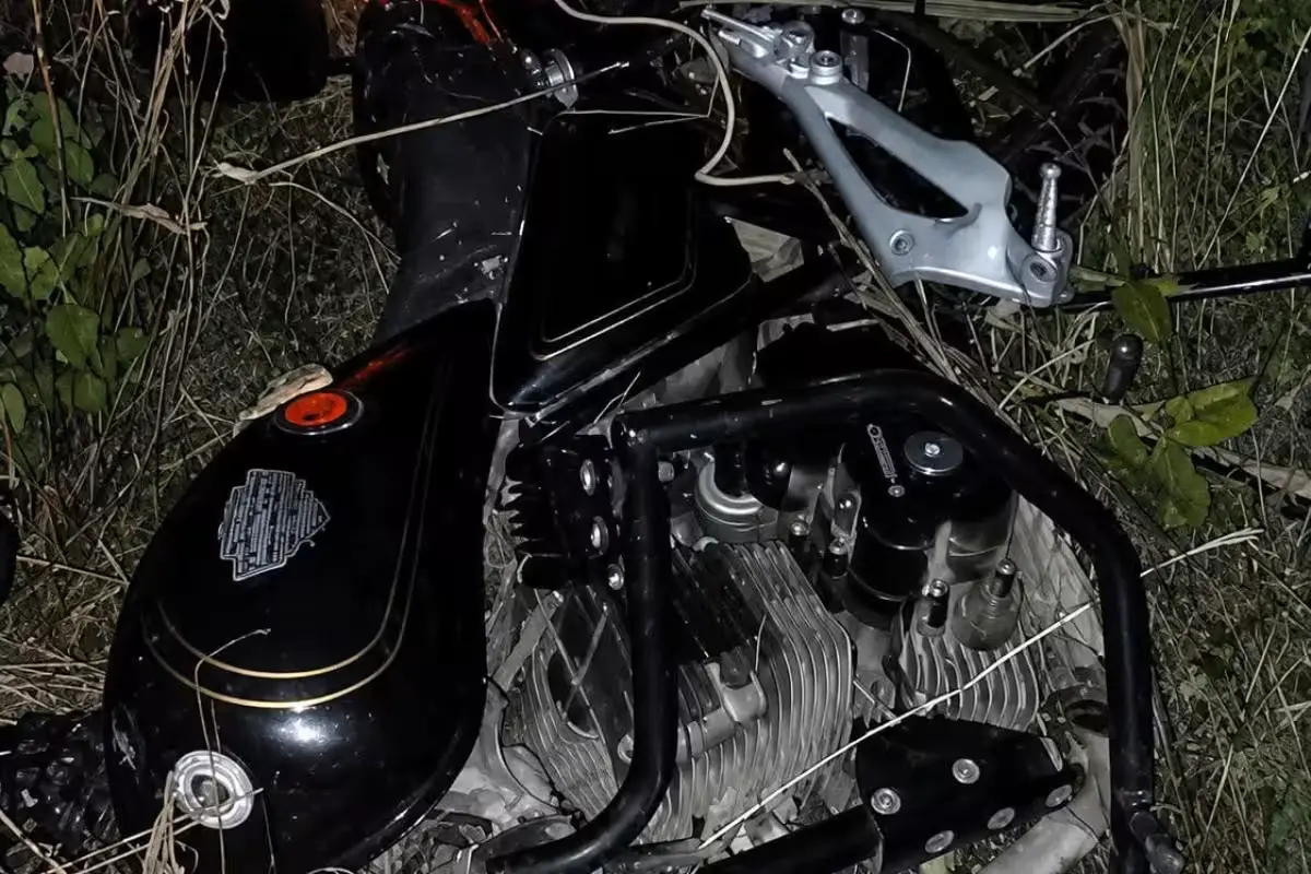 A motorcycle lying on its side in vegetation beside a road at night, with headlights on.