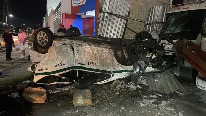 A white taxi lies overturned on a street in Cancun after crashing into poles, with debris scattered around.