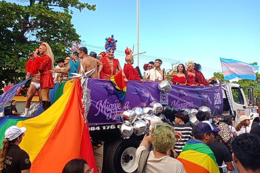 A large crowd of Pride marchers with rainbow flags and banners on a street in Playa del Carmen.