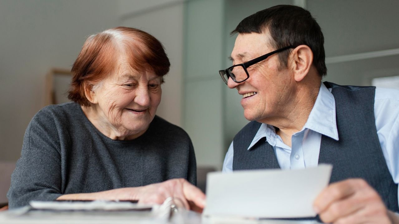 An elderly couple reviewing papers with the IMSS logo in the background