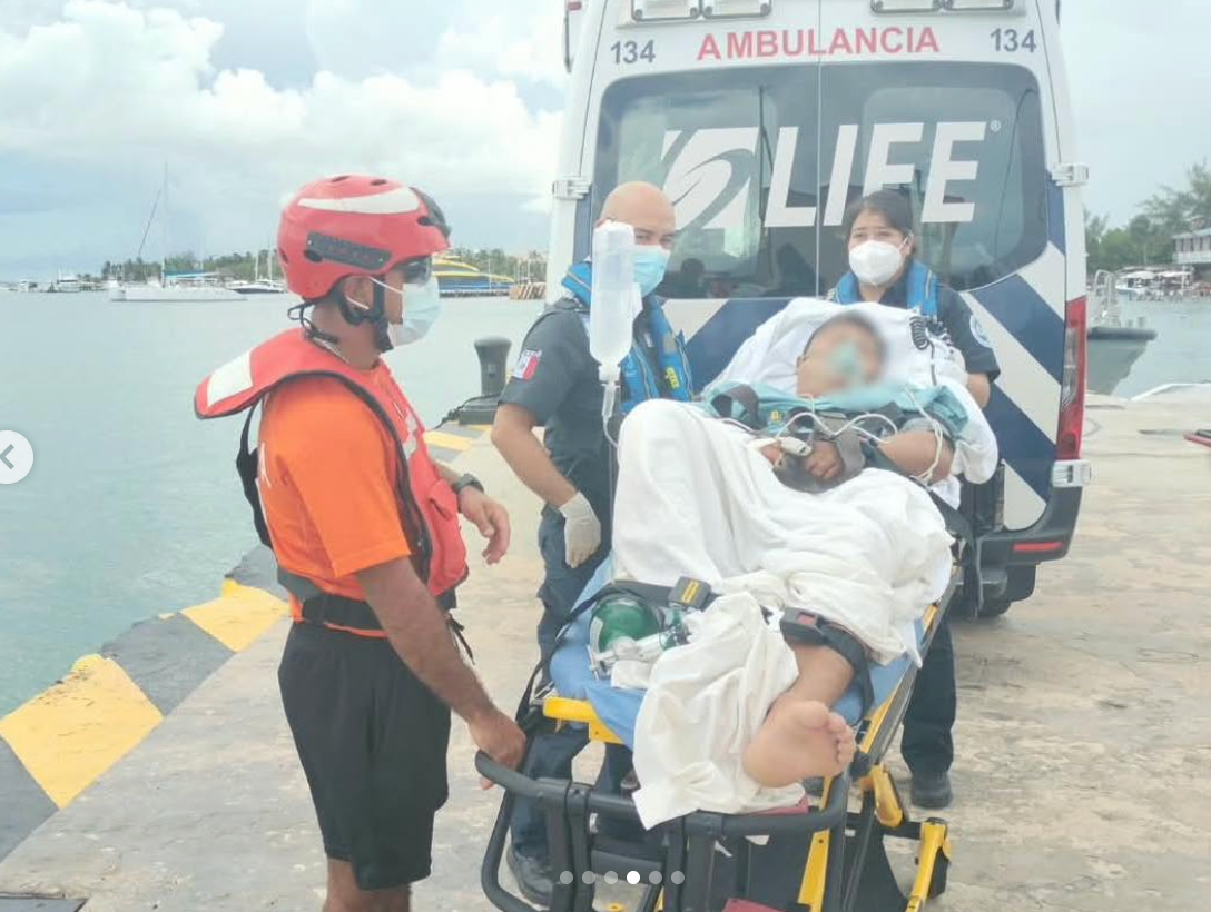 Emergency responders assist a patient on a stretcher near an ambulance at a dock by the water, with boats in the background.
