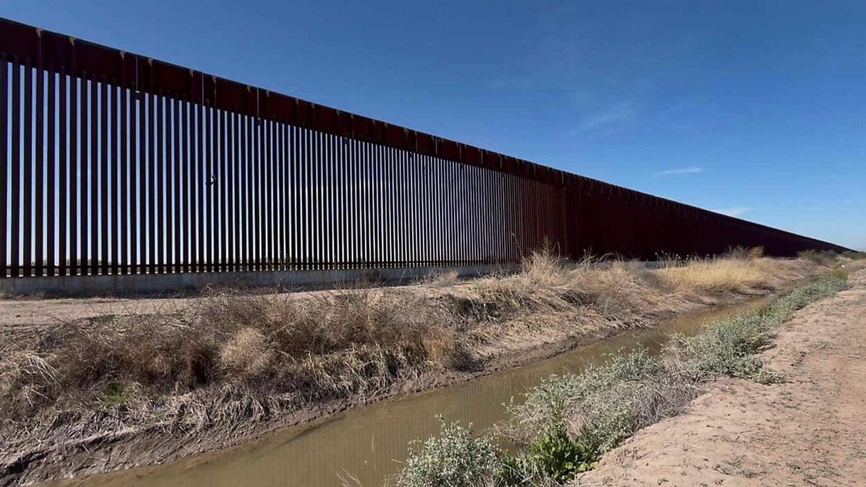 A long section of the U.S.-Mexico border wall with a clear blue sky in the background, surrounded by dry vegetation and a small drainage area.$# CAPTION