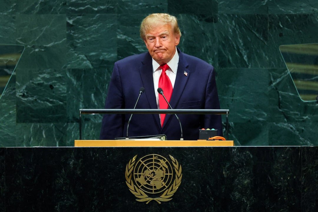 A man in a suit stands at a podium, looking contemplative while addressing a gathering at the United Nations. A green marble backdrop is visible behind him, along with the UN emblem on the podium.