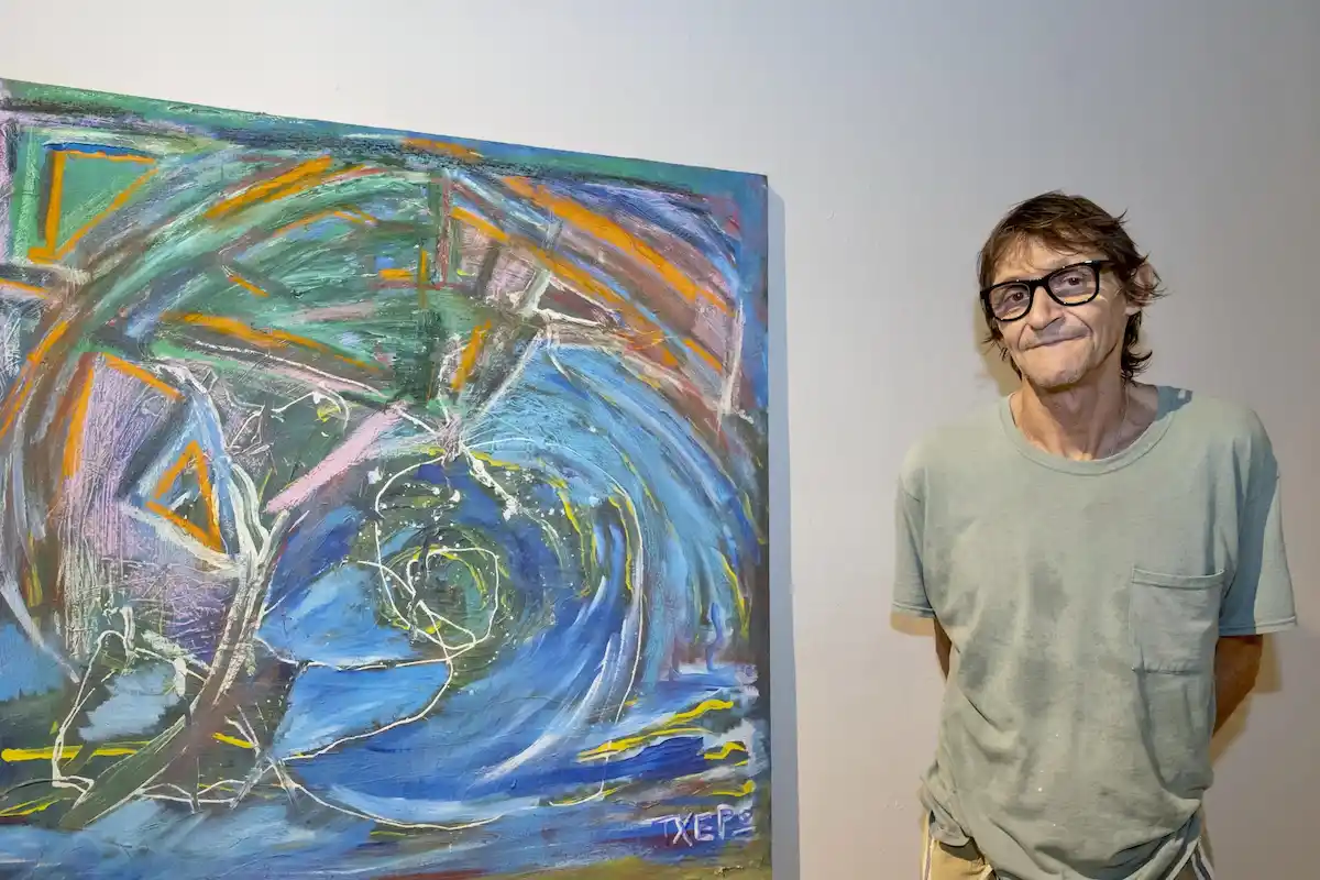 A man stands beside a colorful abstract painting on a wall, smiling and wearing glasses.