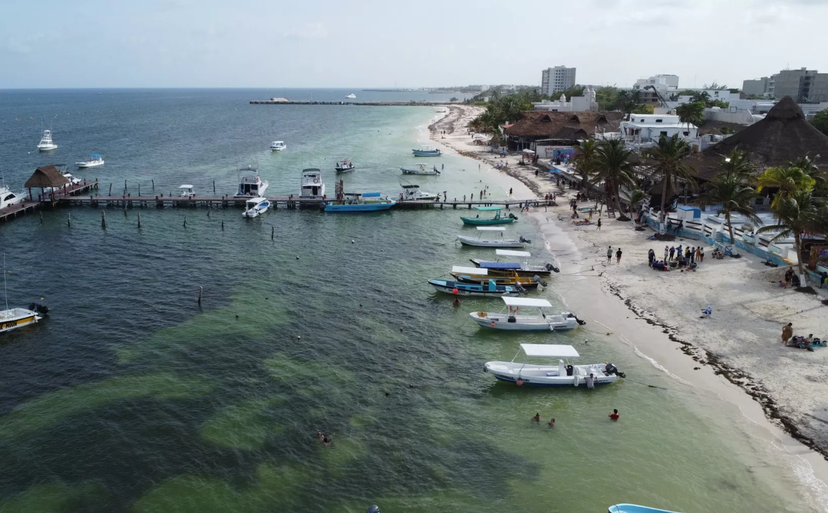 An aerial view shows a sandy beach lined with palm trees and buildings, a wooden pier extending into the water, and numerous small boats moored along the shore and in the turquoise sea.