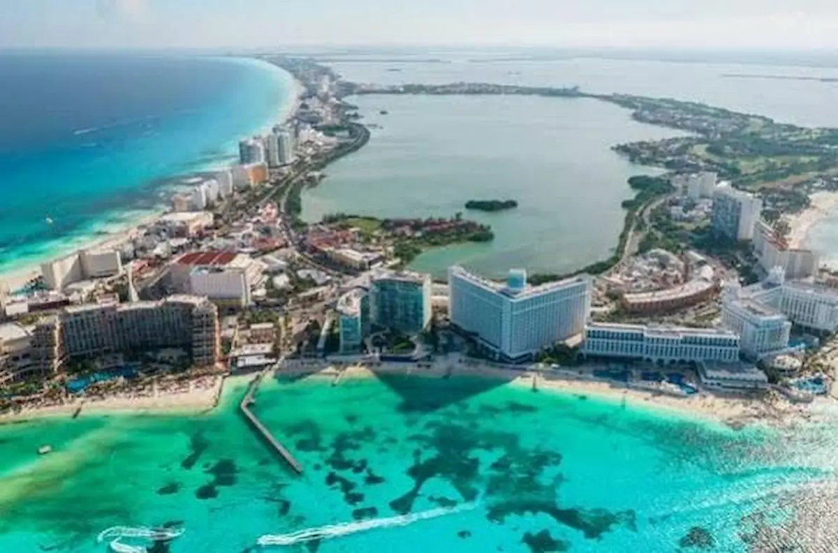 An aerial view shows a narrow strip of beach lined with large hotels between the turquoise Caribbean Sea and a large inland lagoon.