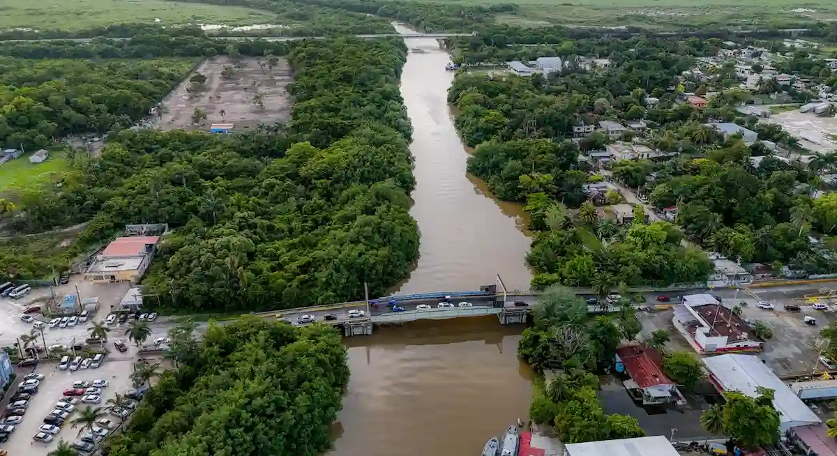 An aerial view shows a wide brown river flowing between dense green vegetation, crossed by a bridge carrying vehicles, with a parking lot and buildings on the left bank and a town on the right bank.