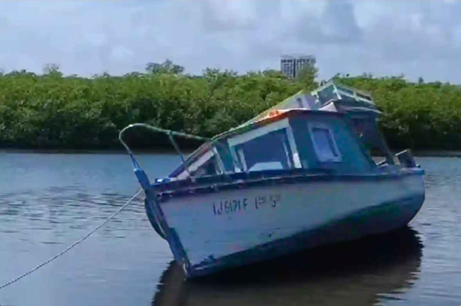 A half-sunken blue boat tethered to the shore in calm waters, surrounded by greenery and distant structures in the background.$# CAPTION