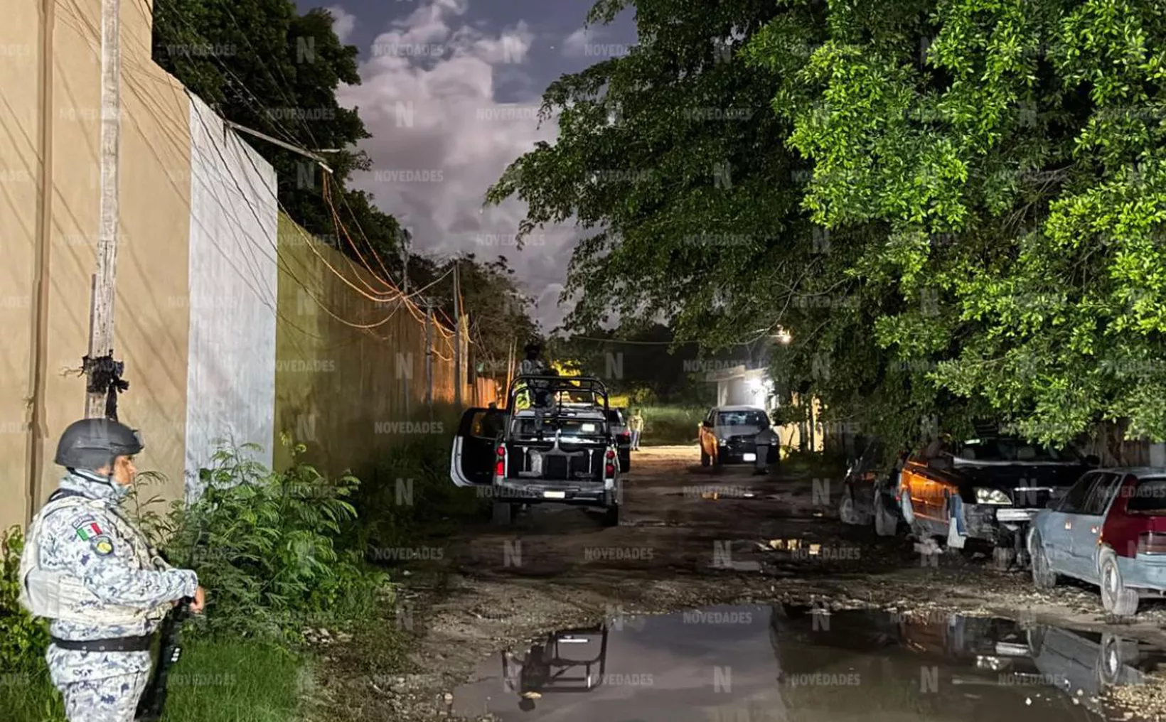 A person in camouflage uniform and black helmet stands holding a rifle on the left of a muddy street lined with parked vehicles, a wall, and trees at night.