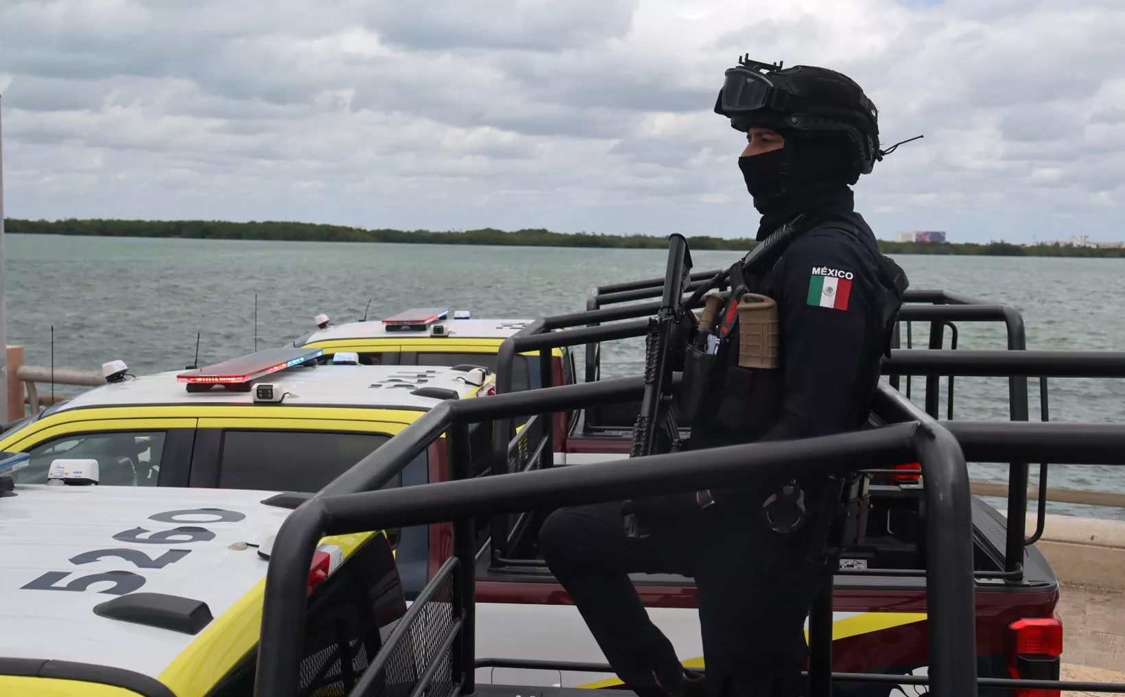 A person in black tactical gear with a helmet, face covering and a Mexico flag patch stands holding a rifle in the bed of a pickup truck behind two white and yellow patrol vehicles, with water and a tree lined shore in the background.