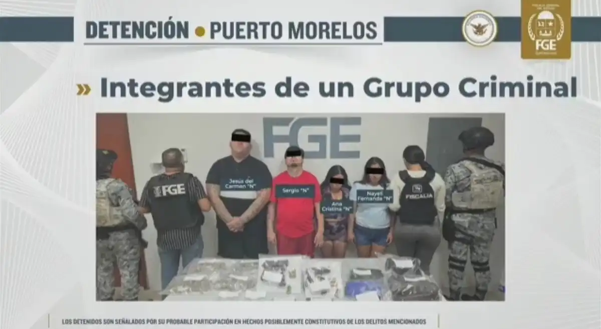 A group of individuals identified as members of a criminal organization, along with law enforcement personnel, during an arrest in Puerto Morelos. Various items are displayed on a table in front of them.$# CAPTION