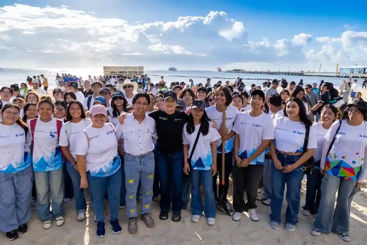 A large group of people wearing white and blue shirts pose together on a sandy beach under a partly cloudy sky, with a sign reading Mega Limpieza Costera 2025 in the background.