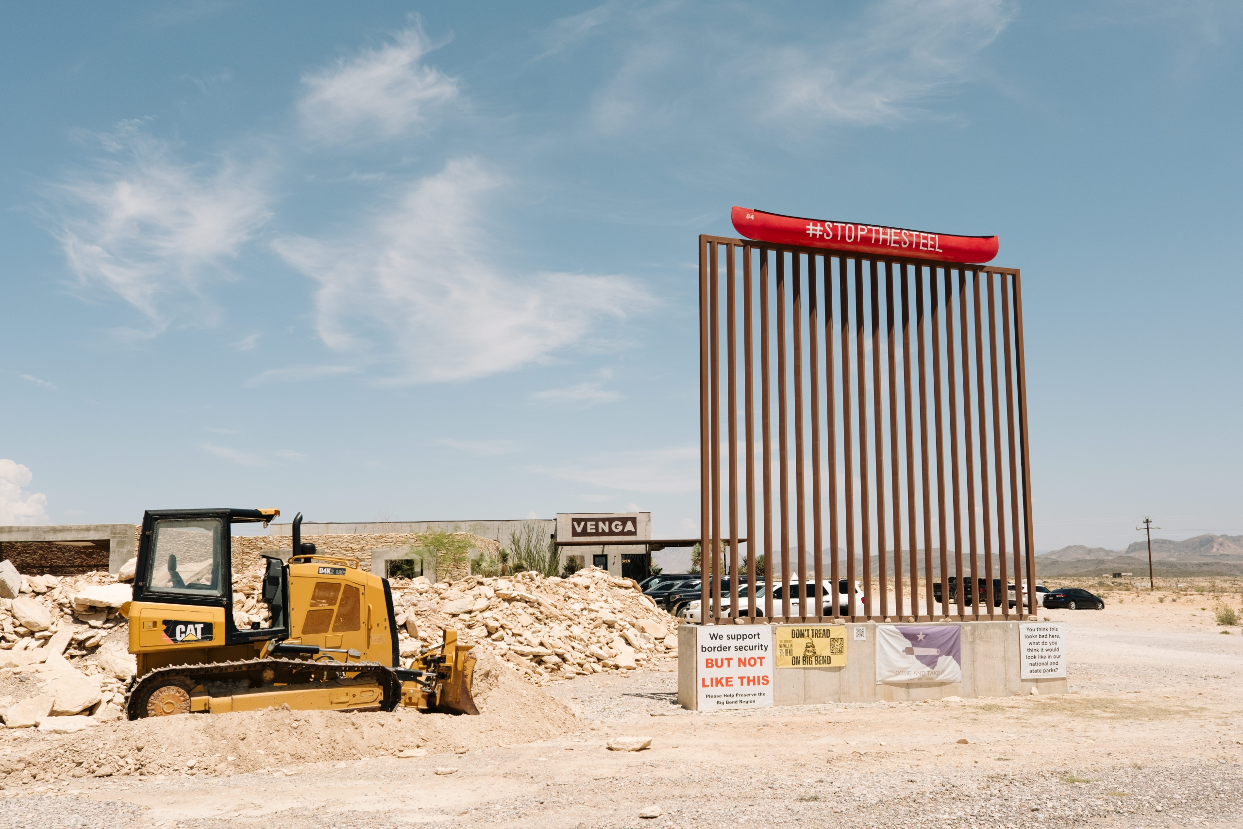 A bulldozer beside a modern art installation with a red canoe at the top, displaying signs related to border security and protest messages in a desert-like setting.$#$ CAPTION