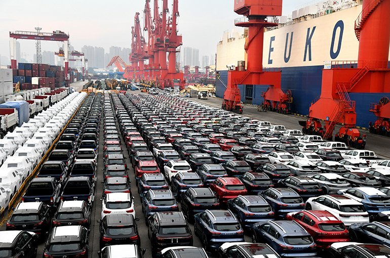 A large shipment of parked cars at a shipping dock with cranes in the background and containers on the left.$#$ CAPTION