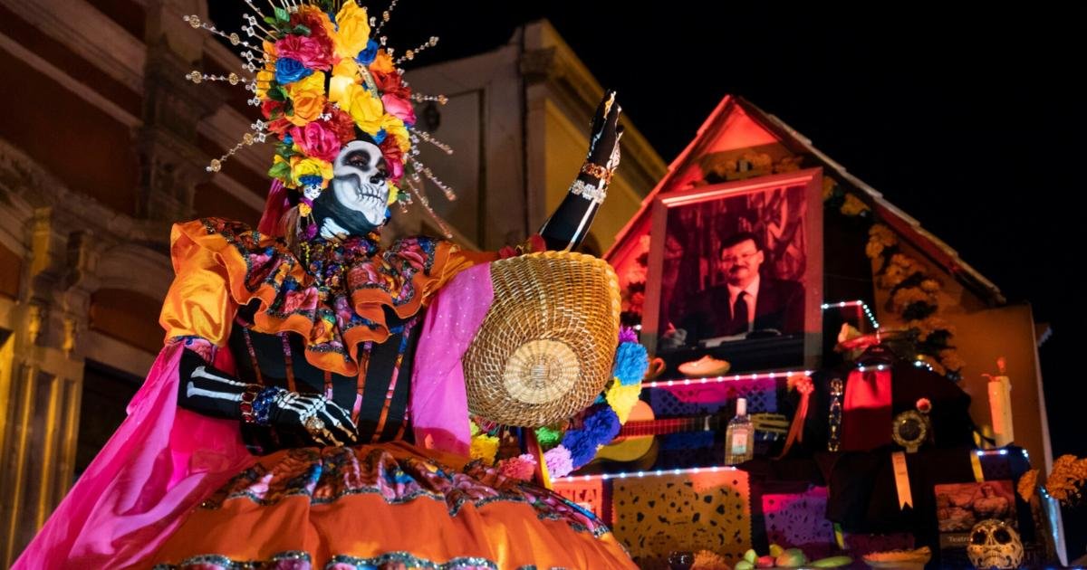 A person in a Catrina costume with a skull face painted and a large colorful floral headdress stands with one arm raised in front of an illuminated altar display at night.