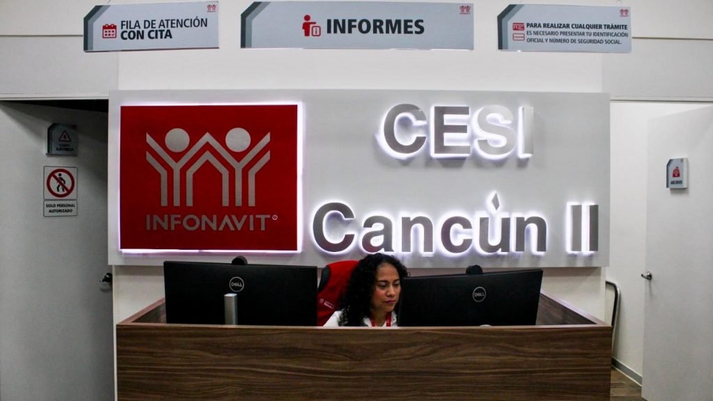 A woman sits behind a wooden counter with two Dell monitors beneath a large illuminated sign reading CESI Cancun II and an Infonavit logo, with overhead signs for appointments and information.