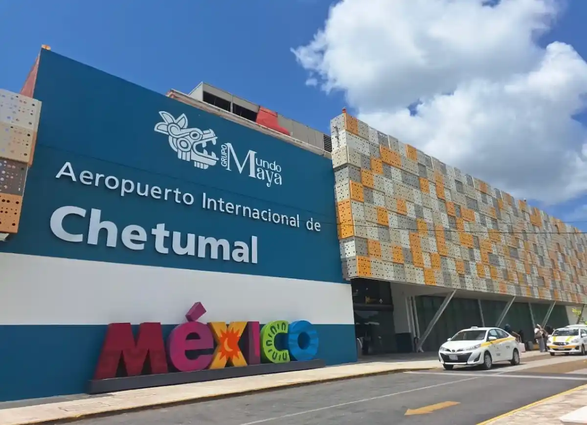 The exterior view of Chetumal International Airport with a modern design and colorful lettering for "México" displayed prominently. The sky is clear with a few clouds.
