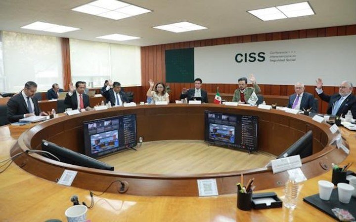 A group of people seated around a large circular wooden conference table in a room with a CISS sign on the wall, several of them raising their hands.