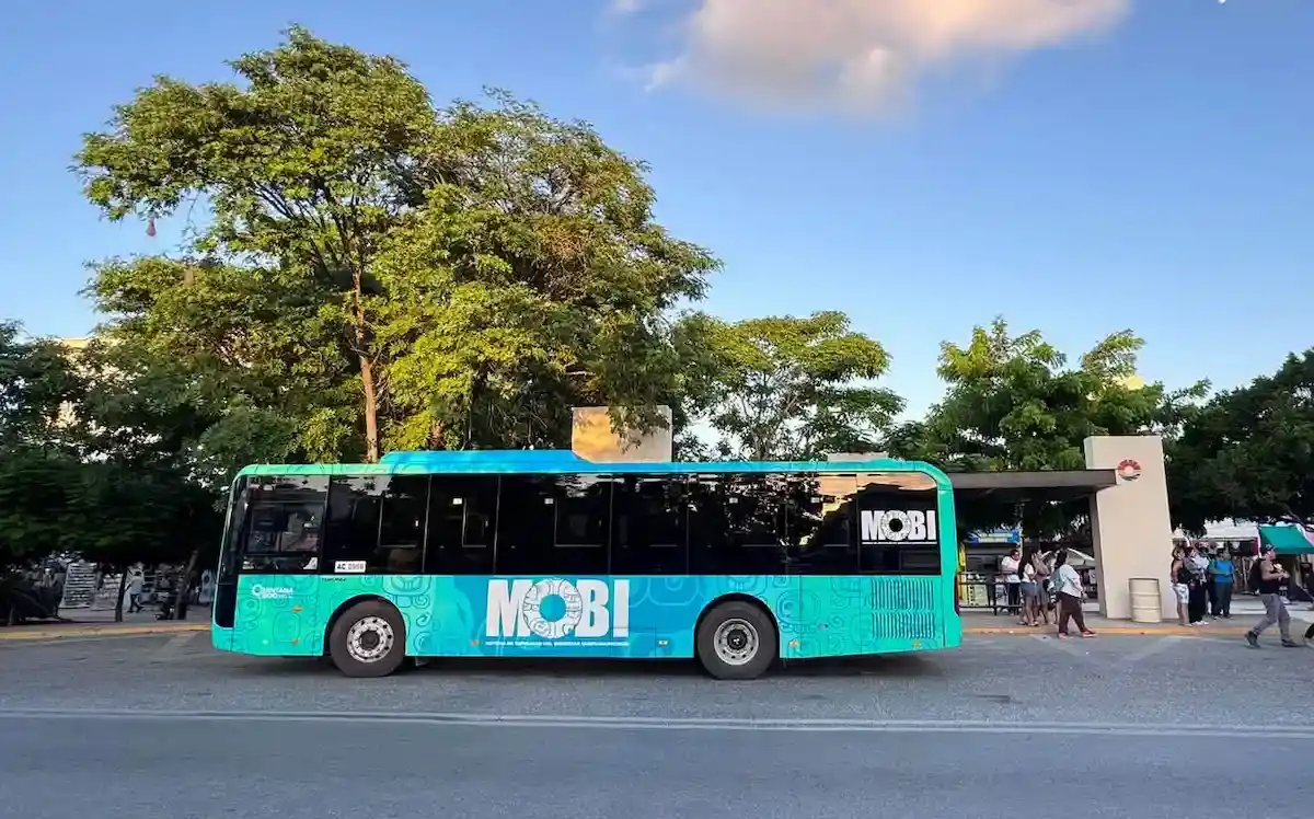 A turquoise city bus with the name "MOBI" parked at a transit stop, surrounded by trees and people waiting at the station.$#$ CAPTION