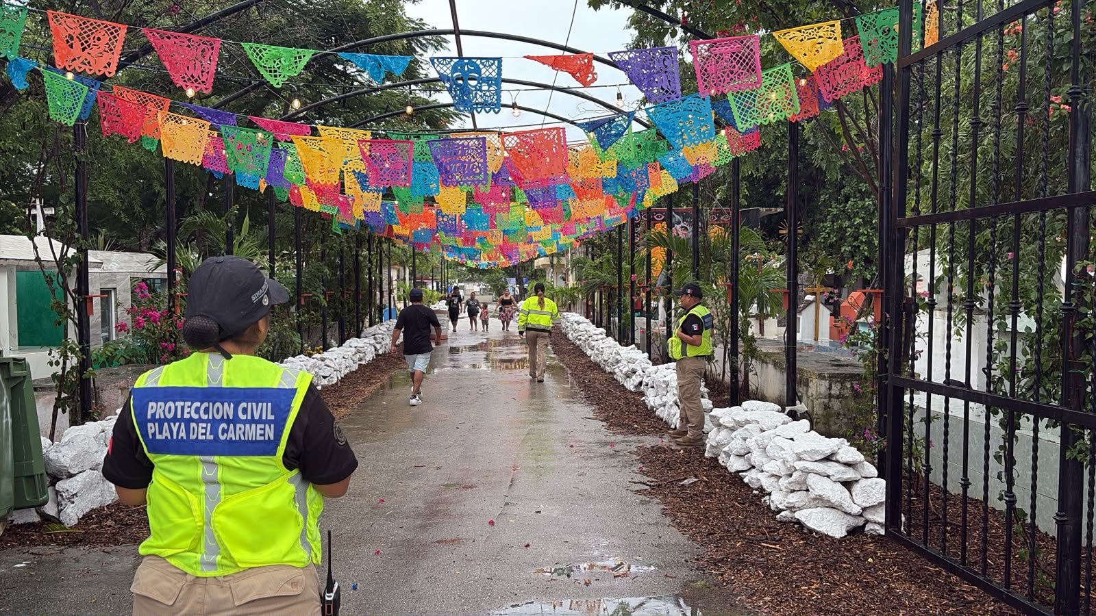 Two civil protection officers in yellow vests stand along a wet paved path lined with white stones and strings of colorful papel picado banners overhead, with people walking in the distance.