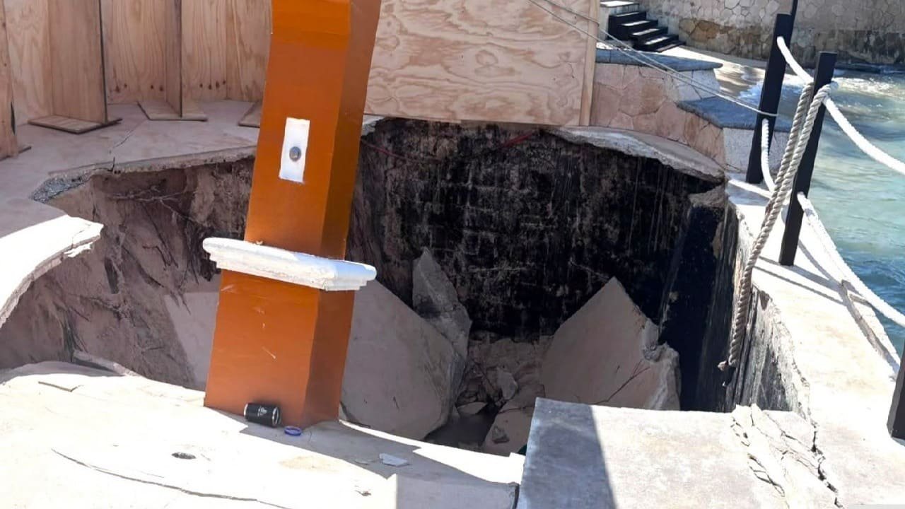 A large hole with broken concrete and dark debris sits beside an orange post and plywood panels near a pool edge.