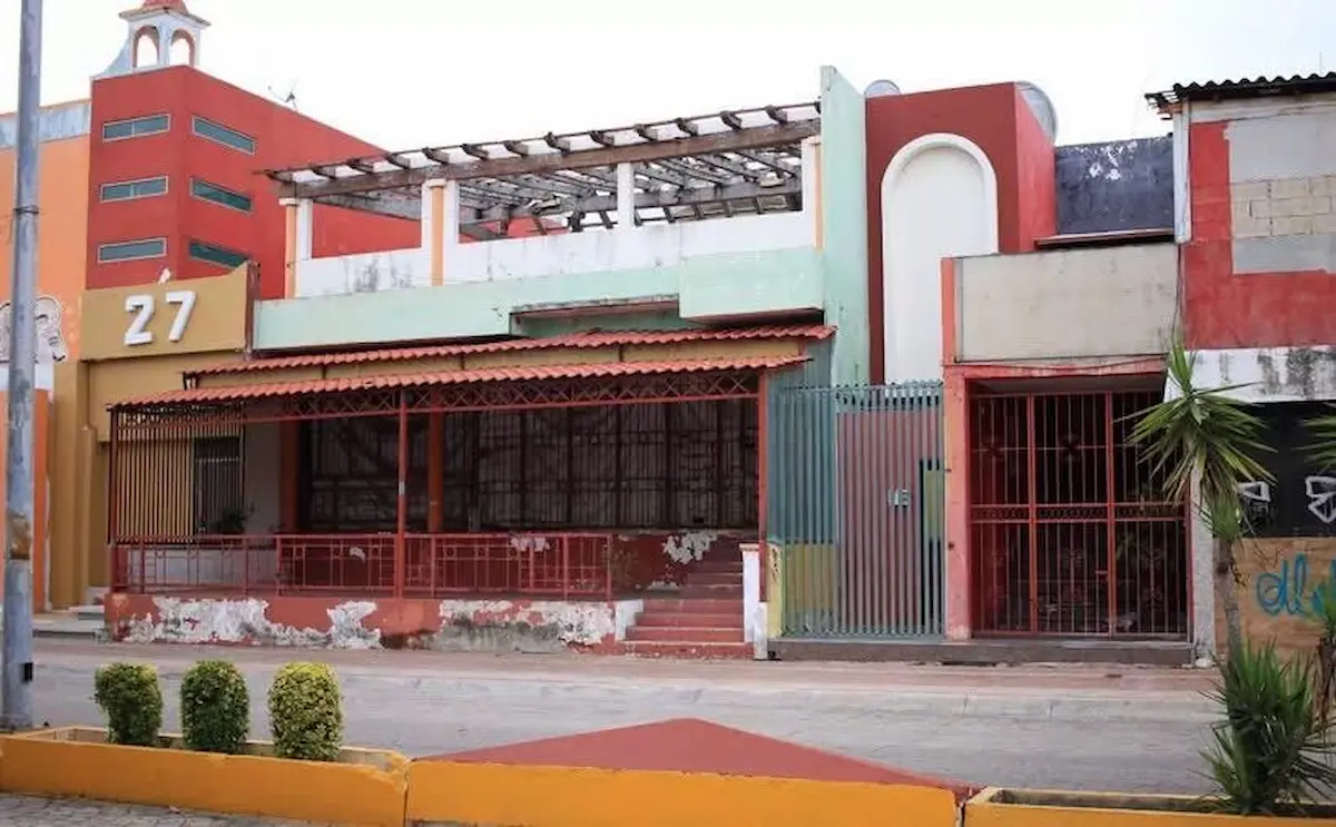 A street view shows a multi colored building with a red and yellow facade bearing the number 27, a covered porch with red metal grilles, a green gate, and a neighboring red building with an arched niche.