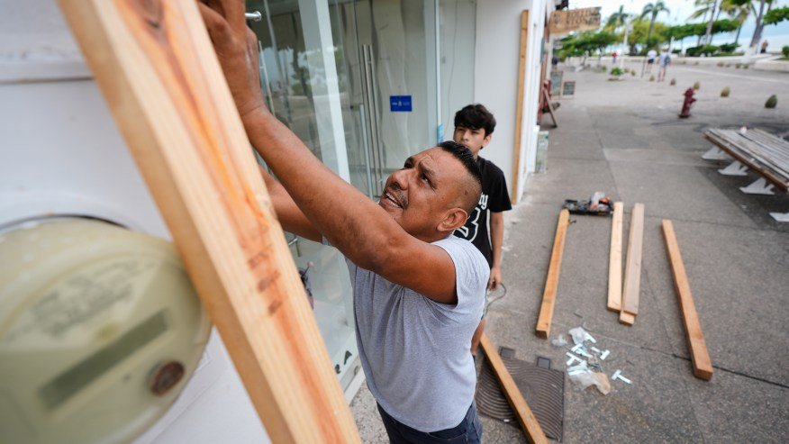 Two men boarding up a storefront in preparation for an approaching storm, with wooden planks and tools visible on the ground.