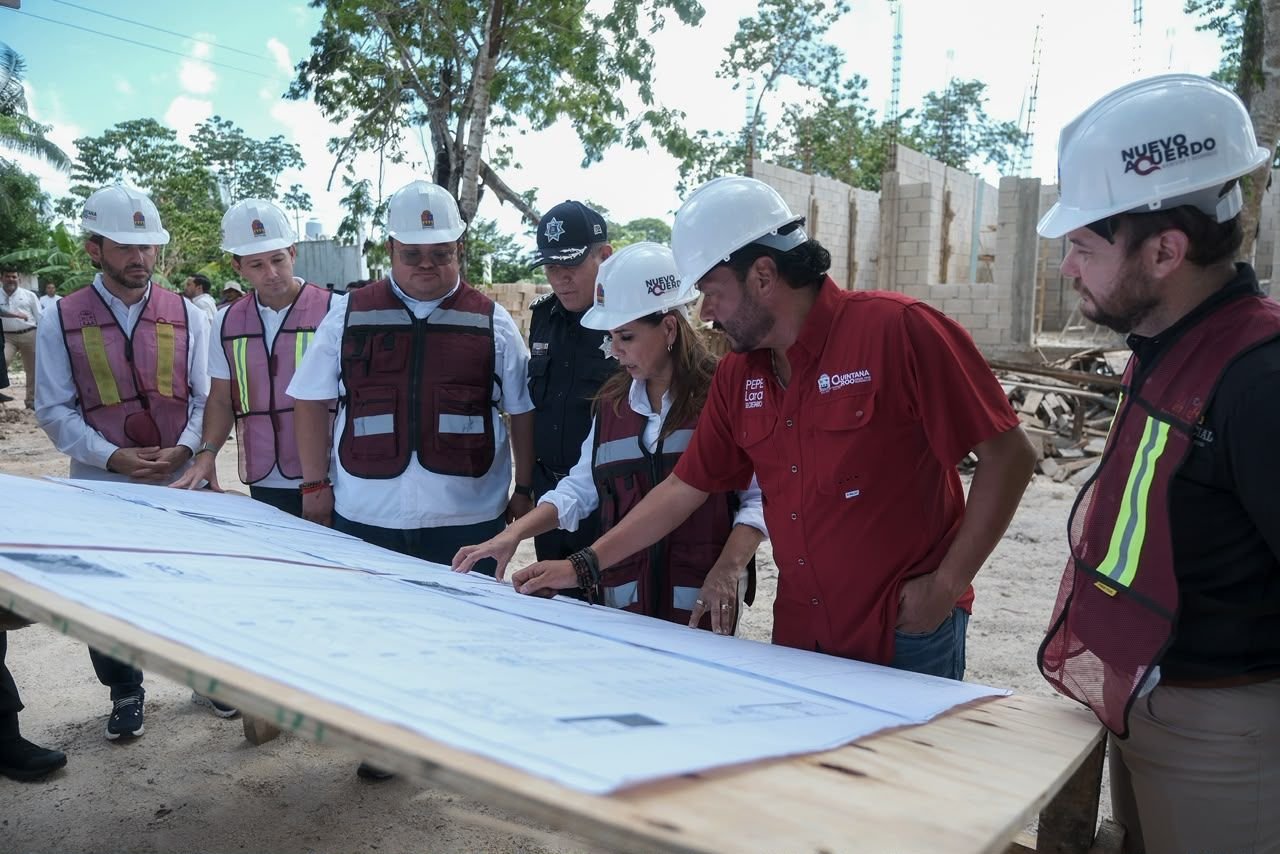 A group of people wearing hard hats and safety vests stand around a wooden table looking at large blueprints at a construction site with unfinished block walls in the background.