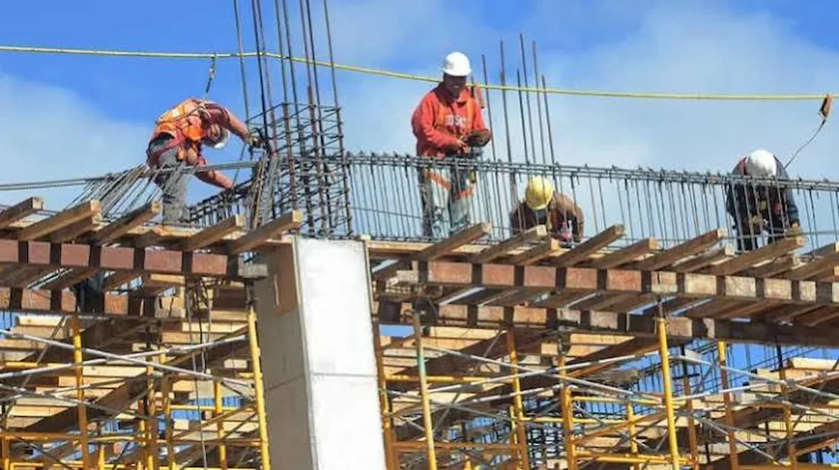Four construction workers on a building site, working on a concrete structure with scaffolding and rebar visible under a blue sky.