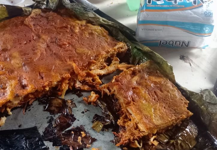 A large piece of reddish brown cooked meat rests on dark banana leaves with a smaller portion cut away, next to a pack of Nubel tissues on a light surface.